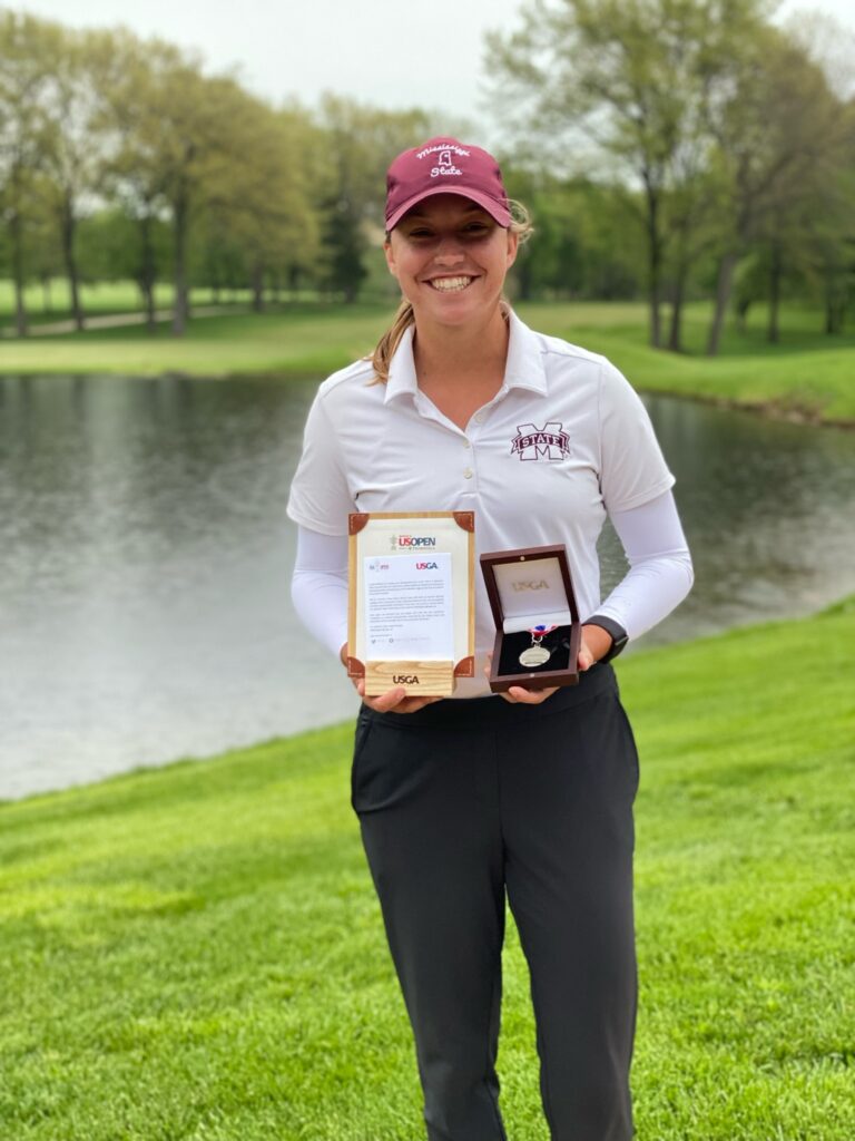 woman standing with awards on a golf course, wearing Mississippi State Clothing.