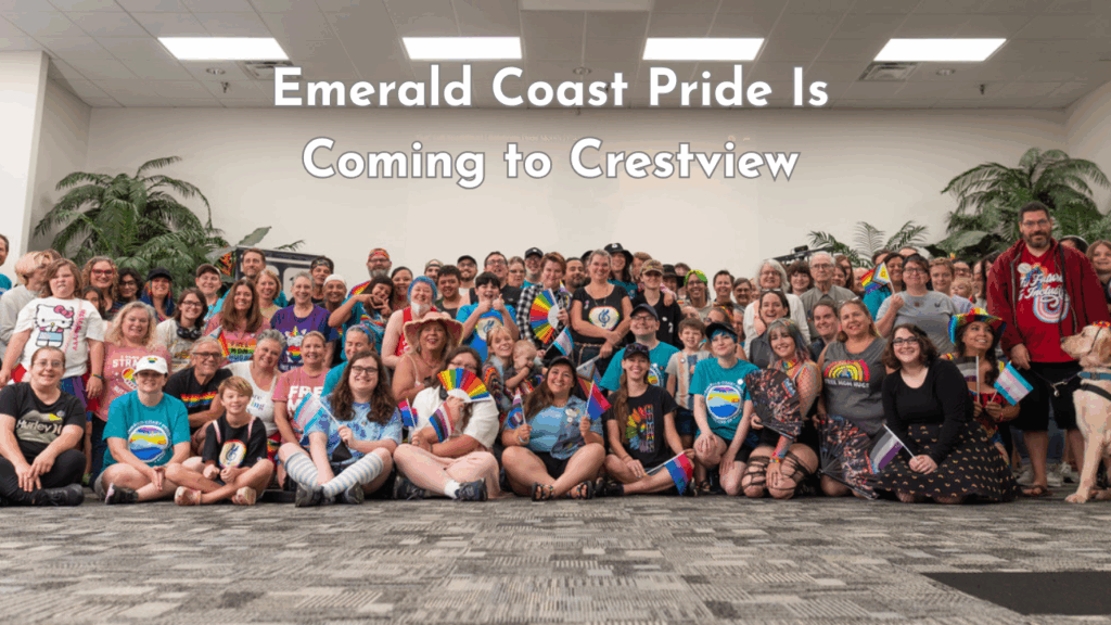 A large group of smiling people of all ages pose indoors at Emerald Coast Pride in Crestview, many holding pride flags and wearing rainbow clothing.