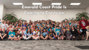 A large group of smiling people of all ages pose indoors at Emerald Coast Pride in Crestview, many holding pride flags and wearing rainbow clothing.
