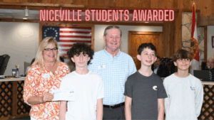 Kristi Evans and Mayor Daniel Henkel pose with three scholarship recipients at Niceville City Hall. Evans holds a ceremonial scholarship check while the students stand in front, smiling. An American flag and the Florida state seal are visible in the background.