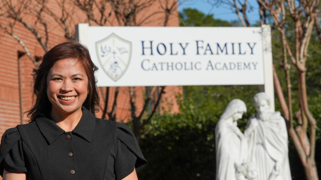 a woman stands in front of a banner that welcomes people to Holy Family Catholic Academy in Niceville Florida.