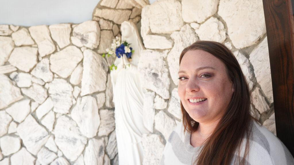 a woman smiles inside a grotto in front of a statue of the Blessed Virgin Mary