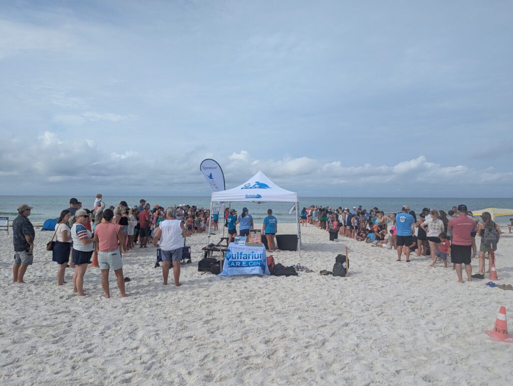 Crowd of people gathered on a sandy beach around white tents with banners, ocean in the background.