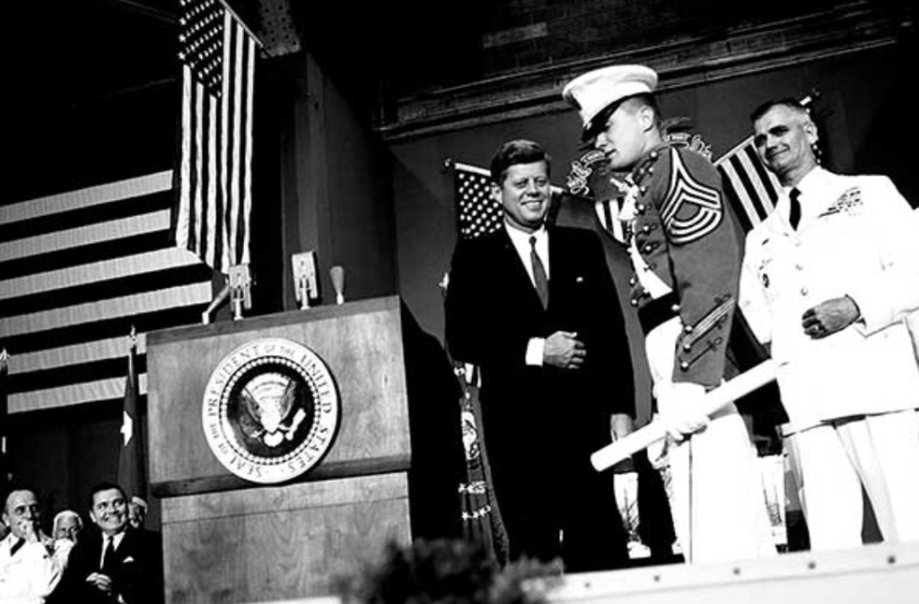 Former President John F. Kennedy on stage with a Marine and a Navy officer during a ceremony, American flags and the presidential seal in the background.