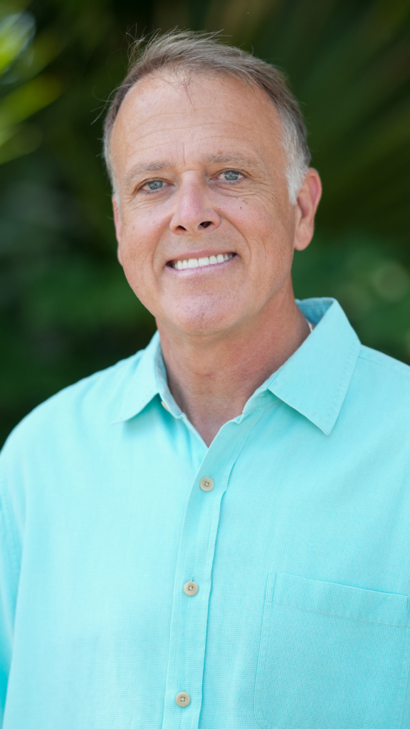 Portrait of a smiling man with short gray hair, wearing a turquoise button-down shirt, outdoors with greenery in the background.