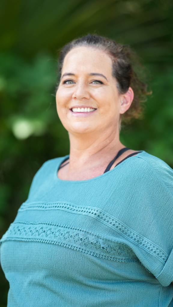 Portrait of a smiling woman outdoors, wearing a teal top with a green garden background.
