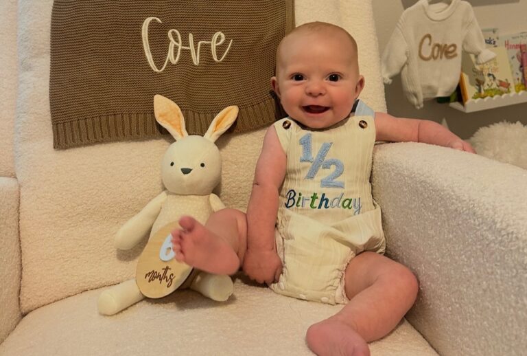 Smiling baby in a light outfit sits on a cream chair with a plush bunny, a brown blanket labeled Core behind them, and a cozy nursery shelf in the background.