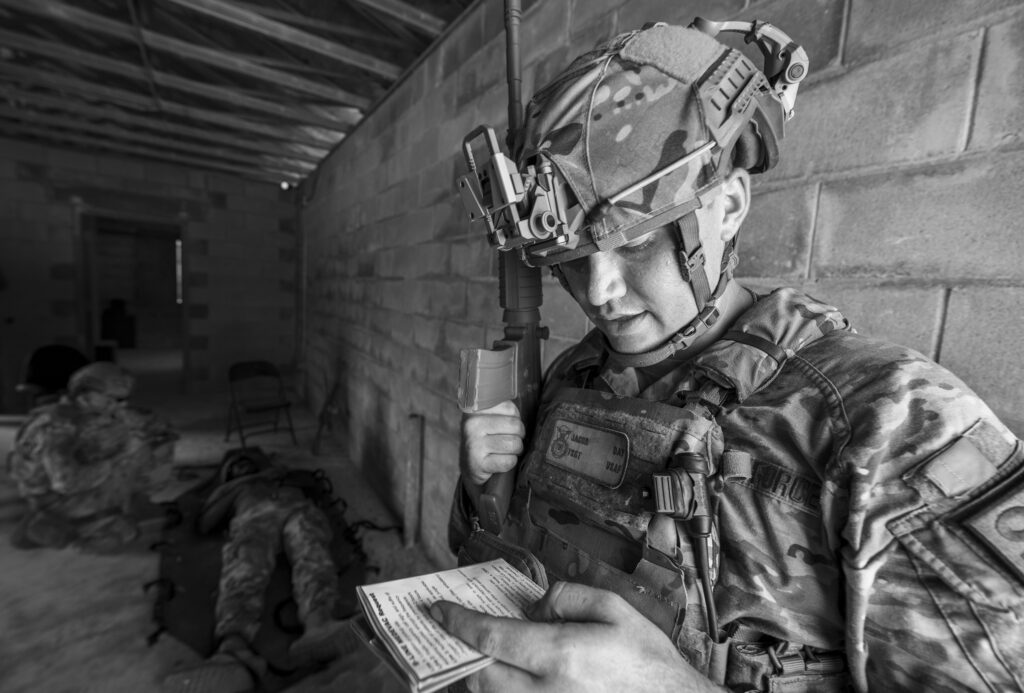 Soldier in camouflage gear and helmet writing on a notepad inside a ruined building.