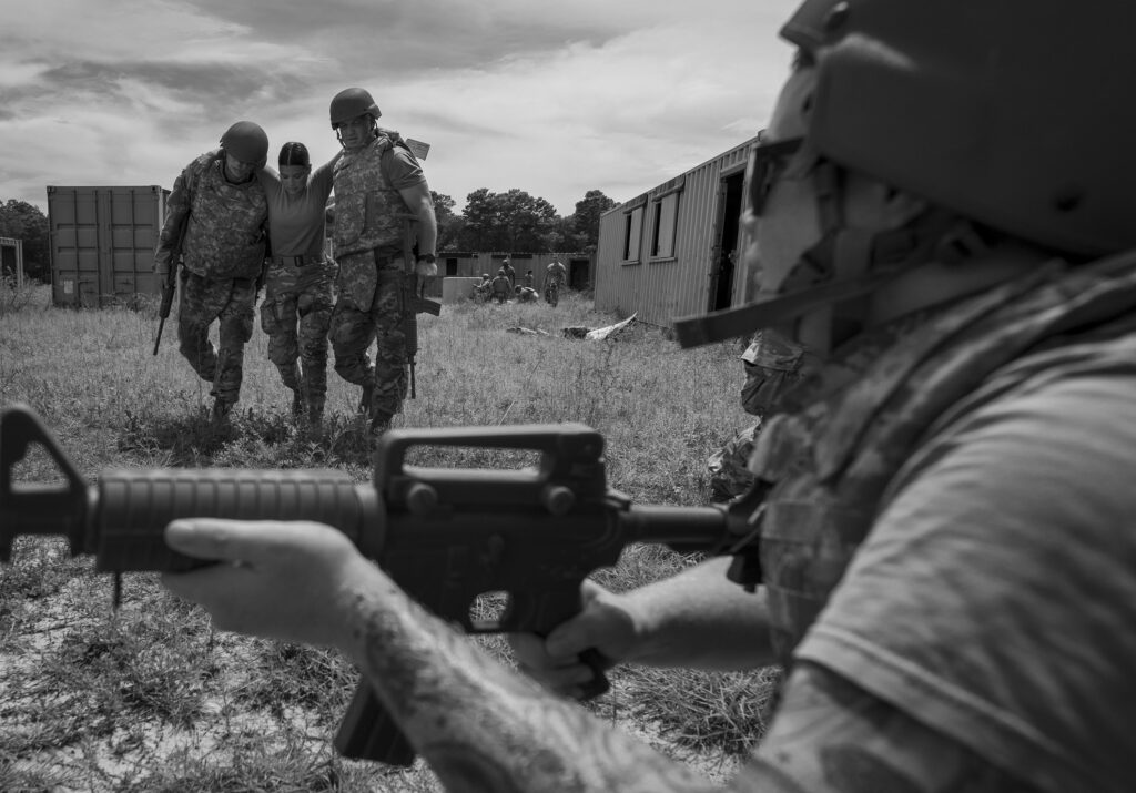 Three soldiers help a wounded teammate walk across a field during training; a fourth soldier aims a rifle in the foreground.