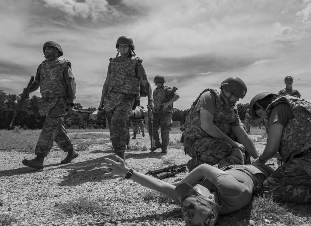 Soldiers in camouflage uniforms and helmets stand and kneel in a rocky outdoor training area with rifles at the ready.