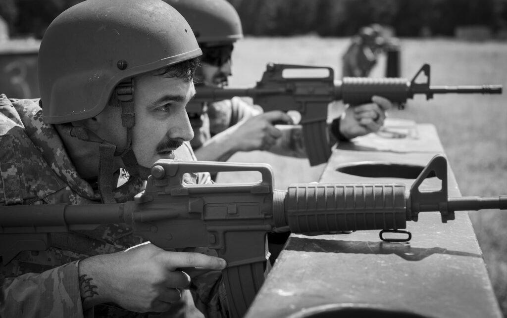 Soldier in camouflage and helmet aiming a rifle from a firing position at a range (black and white).