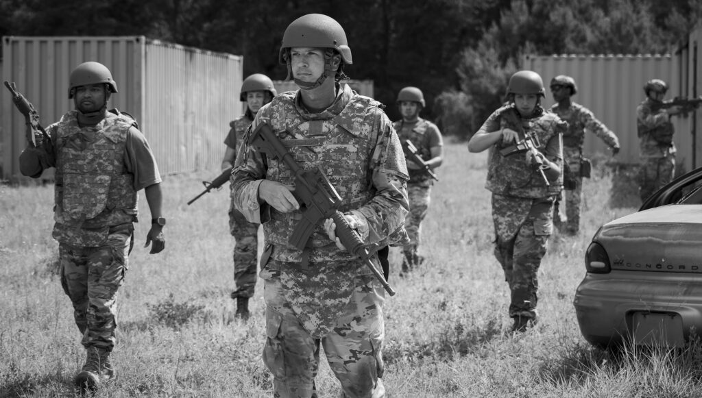 Group of soldiers in camouflage uniforms march with rifles in a grassy outdoor area, near a car and shipping containers in the background.