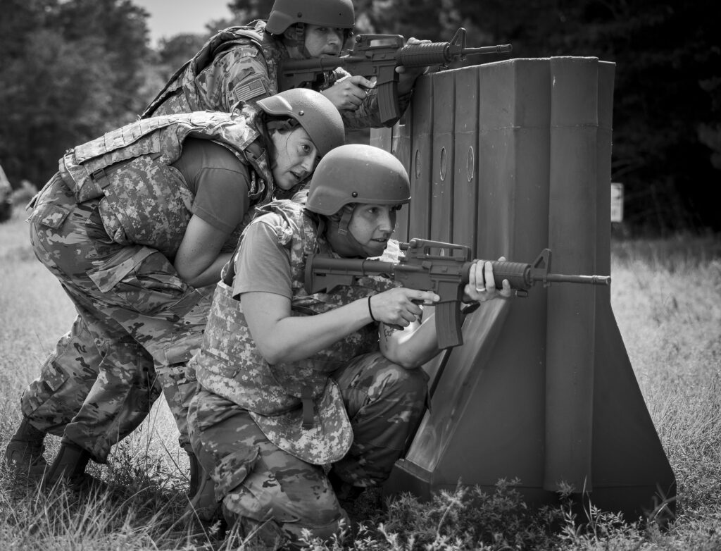 Soldiers in camouflage gear crouch behind a barricade, aiming rifles during a training exercise in a grassy field.