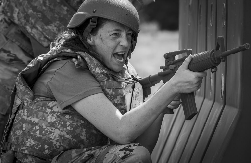 Female soldier in camouflage uniform and helmet aiming a rifle at a metal wall or barrier.