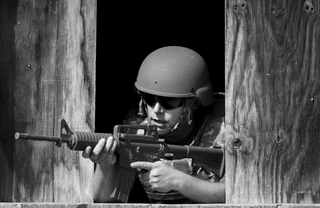 Soldier wearing helmet and tactical gear aims an assault rifle through a wooden barrier opening, in black and white.