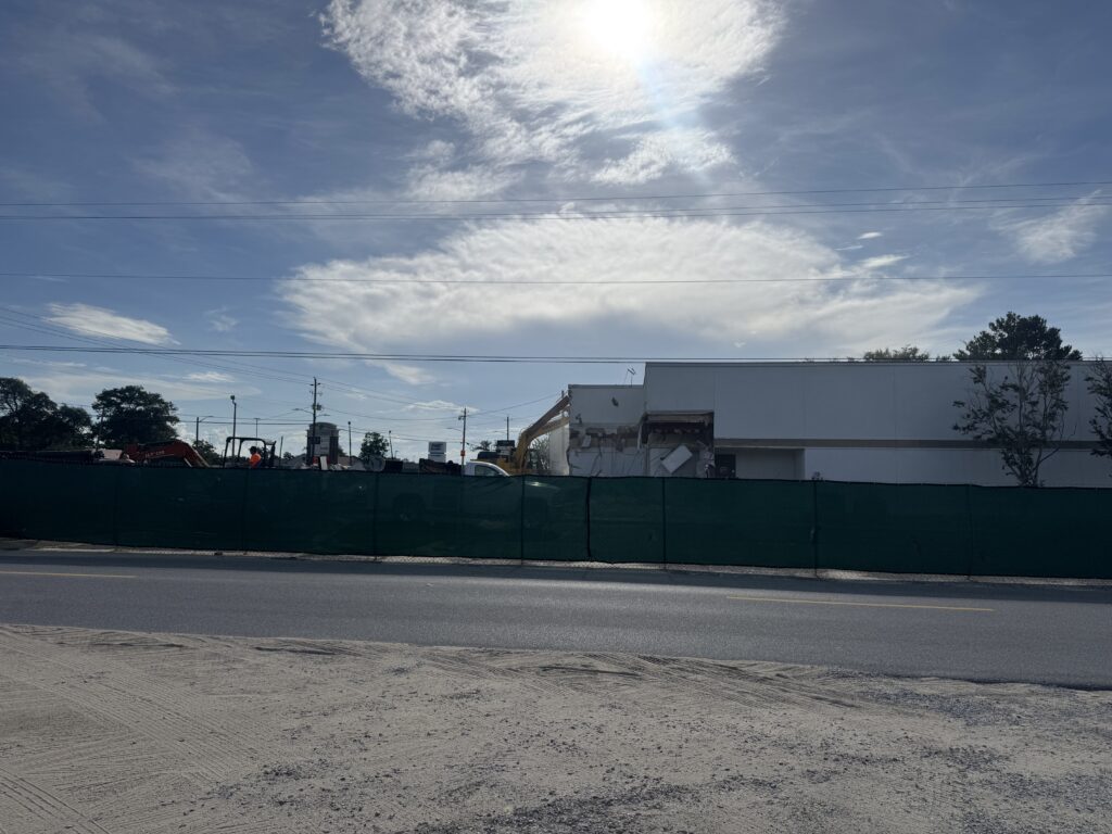 Construction site behind a tall green privacy fence with heavy machinery and a worker, under a bright blue sky and few clouds.