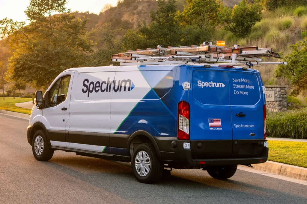 Spectrum service van with ladder rack on roof, parked on a residential street near a grassy hill and trees at sunset.