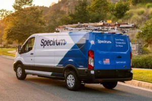 Spectrum service van with ladder rack on roof, parked on a residential street near a grassy hill and trees at sunset.